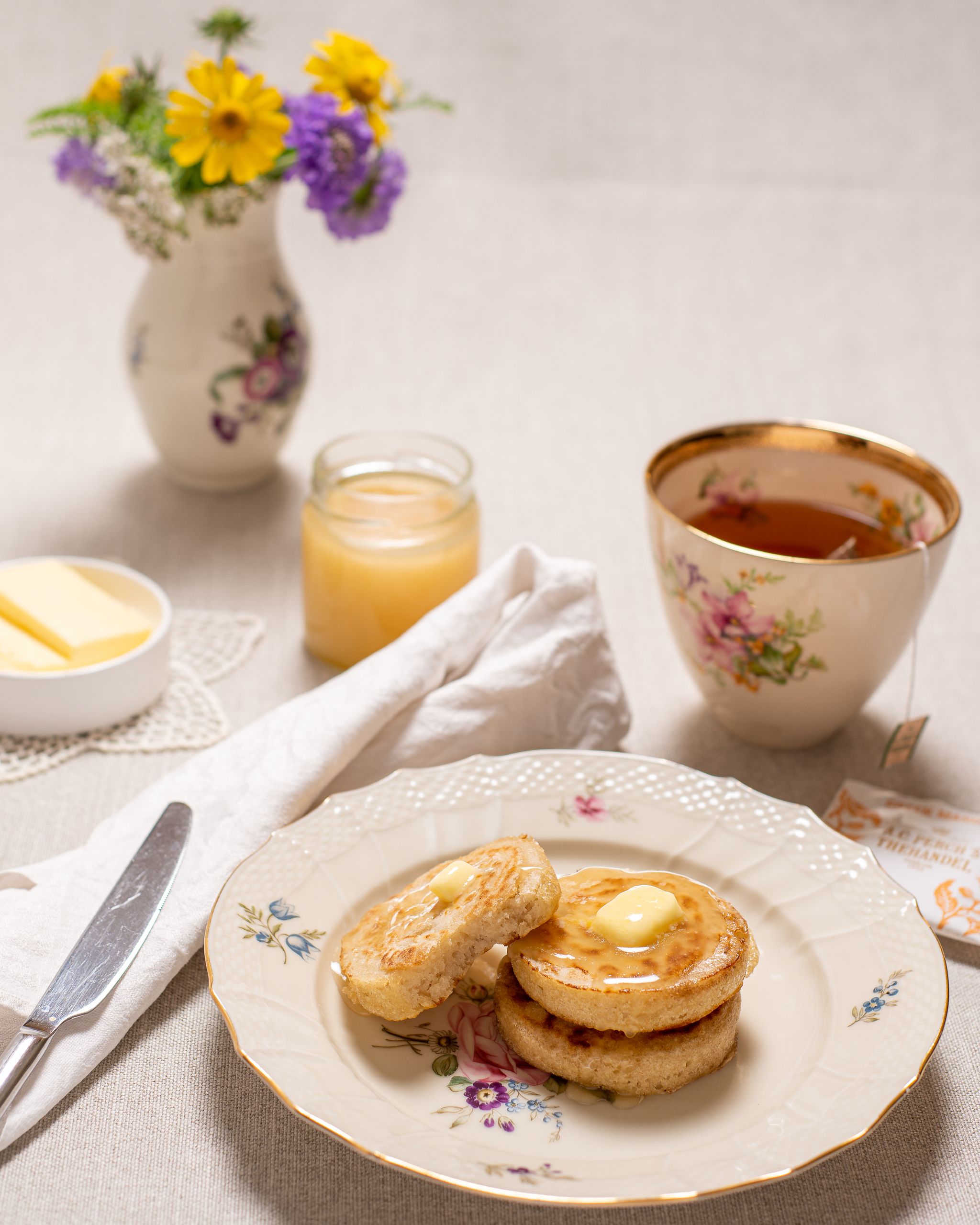 Crumpets anrettet på en tallerken. Omkring tallerkenen er der vase med blomster, krus med the, smør og honning.