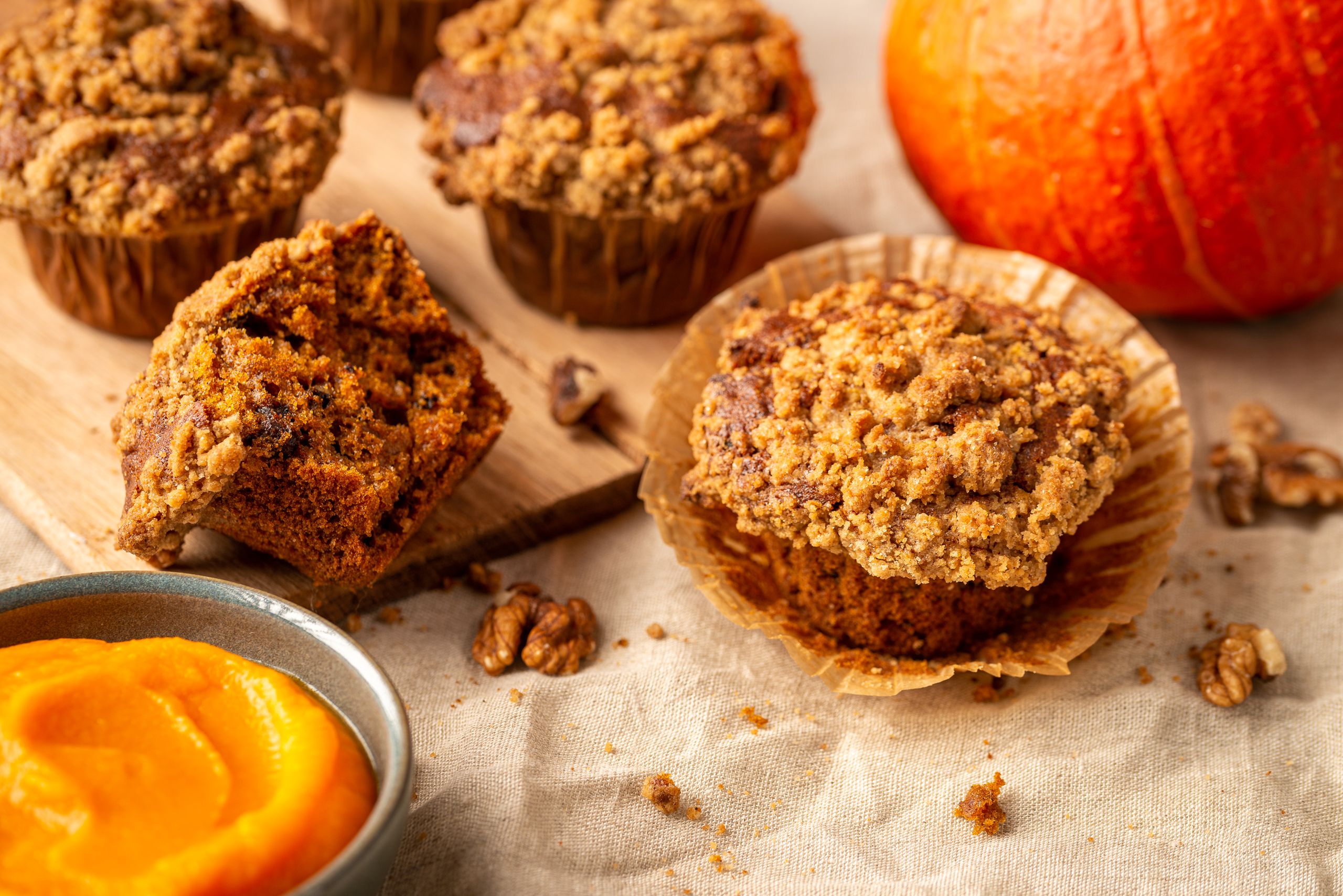 Krydderede græskarmuffins med en sprød Topping. 4 muffins er placeret på en dug. Et orange græskar kan anes i baggrunden. I forgrunden er en lille skål med orange græskarmos.