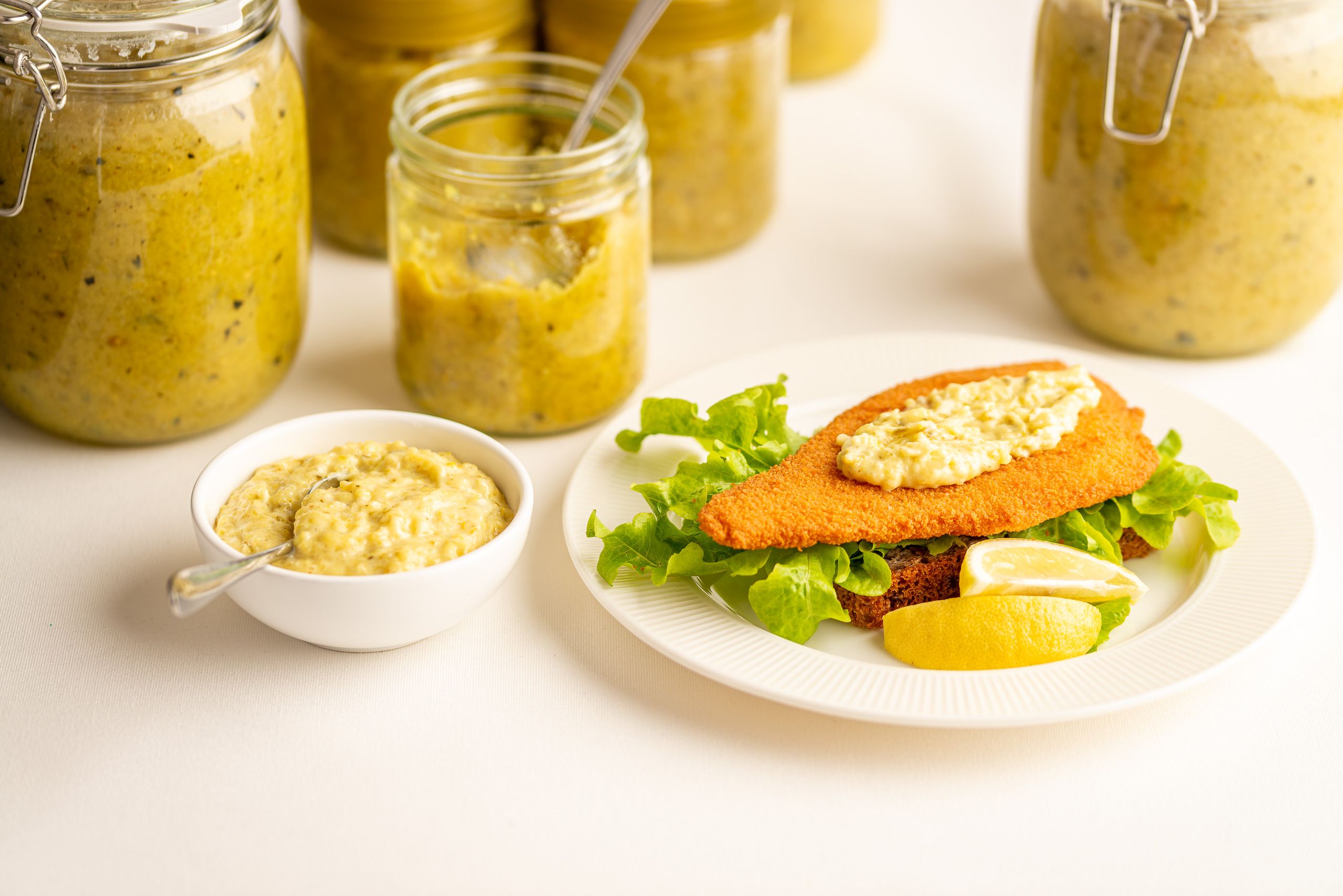 Remoulade relish glas står på en hvid dug. Til venstre i billedet står en lille hvid skål med relish blandet med mayonnaise. I centrum af billedet er en hvid tallerken med en rugbrødsmad med fiskefilet.