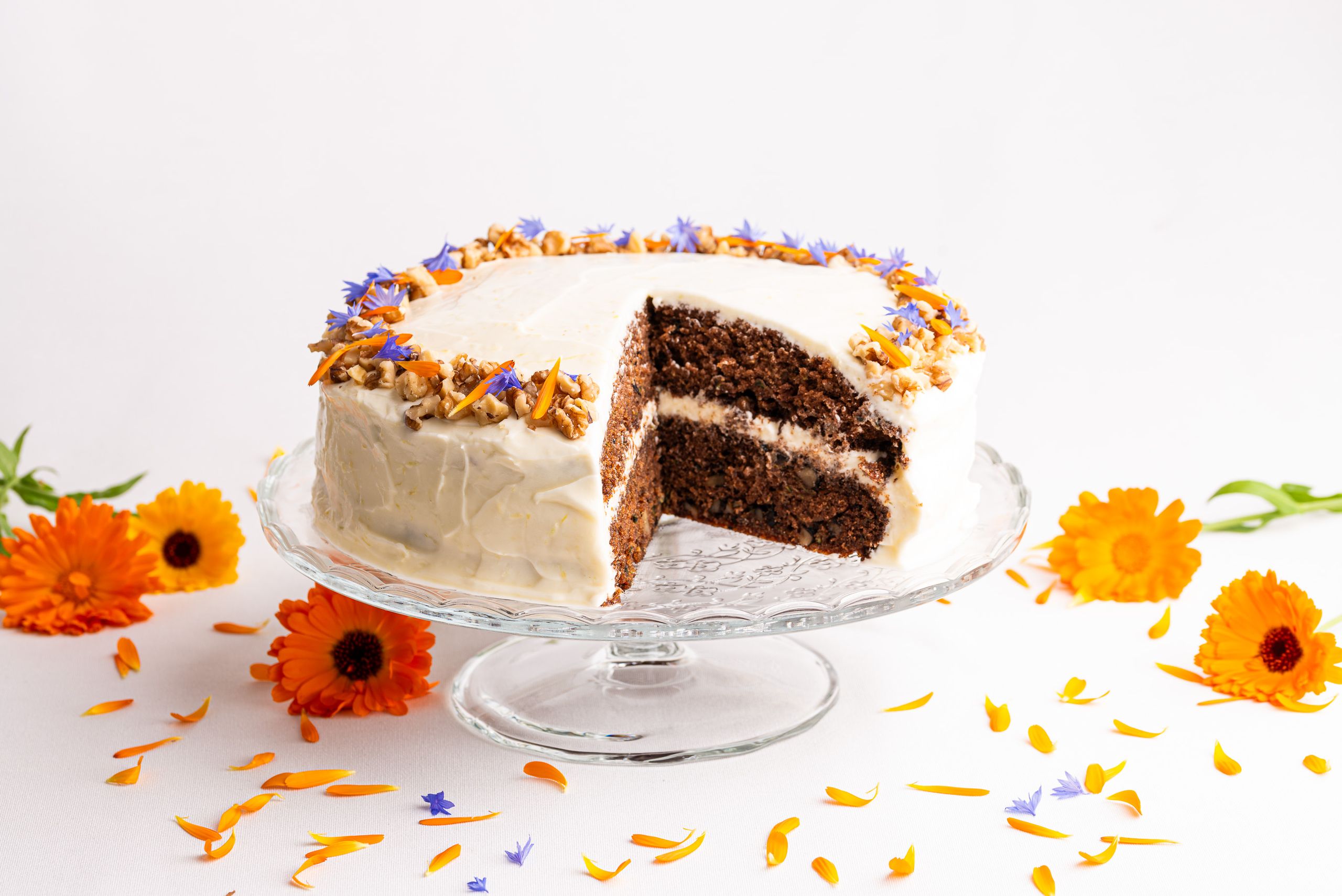 Squashkage med en hvid frosting med flødeost. Pyntet i en cirkel med valnødder og blomsterblade fra orange morgenfruer og blå kornblomster. Kagen ståe på en glasopsats. På bordet under kagen er der spredt blomster hoveder og blade.